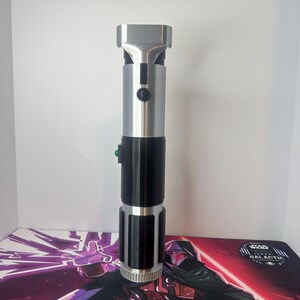 K&ouml;nnte beinhalten: Ein zylindrischer Lichtschwertgriff mit metallisch silbernem Finish an den oberen und unteren Abschnitten, kontrastiert durch einen glatten schwarzen Mittelgriff. Der Griff enth&auml;lt einen kleinen schwarzen runden Aktivierungsknopf und ein gr&uuml;nes Element.