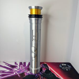 May include: A silver-colored cylindrical lightsaber hilt featuring a bright yellow ribbed emitter section at the top, a black ringed collar, and a central control panel with two circular buttons and two smaller square buttons on the metallic body.