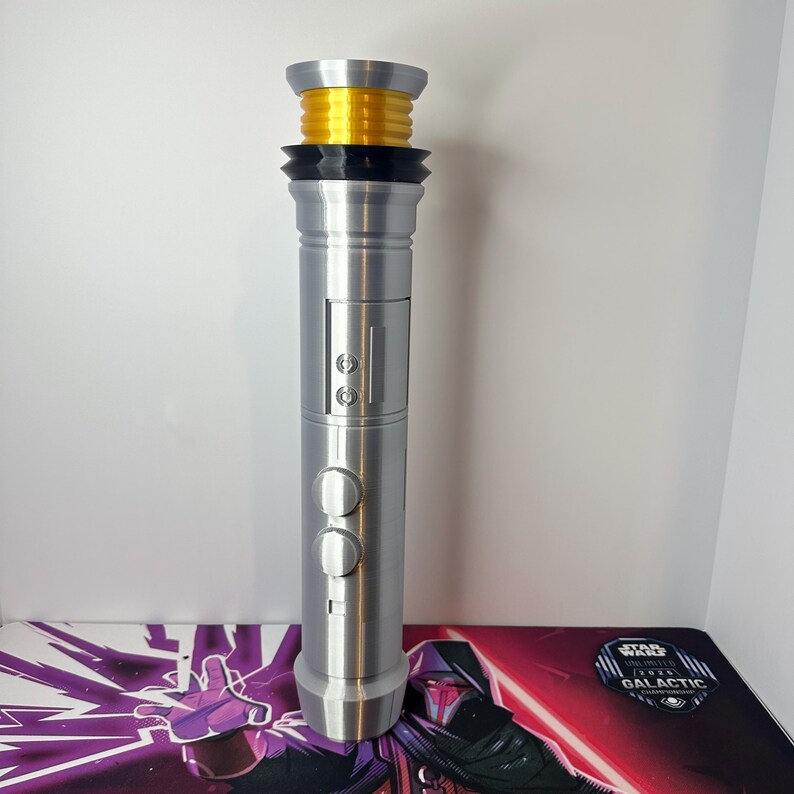 May include: A silver-colored cylindrical lightsaber hilt featuring a bright yellow ribbed emitter section at the top, a black ringed collar, and a central control panel with two circular buttons and two smaller square buttons on the metallic body.