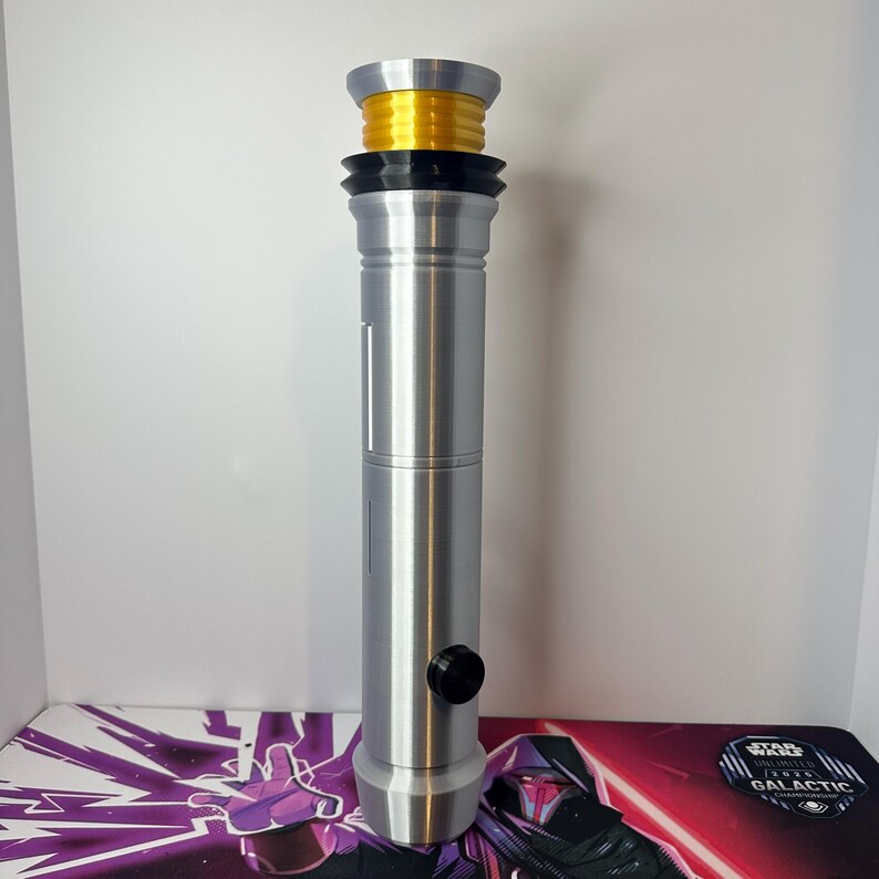 May include: A silver cylindrical lightsaber hilt featuring a bright yellow ribbed emitter section at the top, a black ring detail below the emitter, and a single black circular activation button on the side of the main metallic handle body.