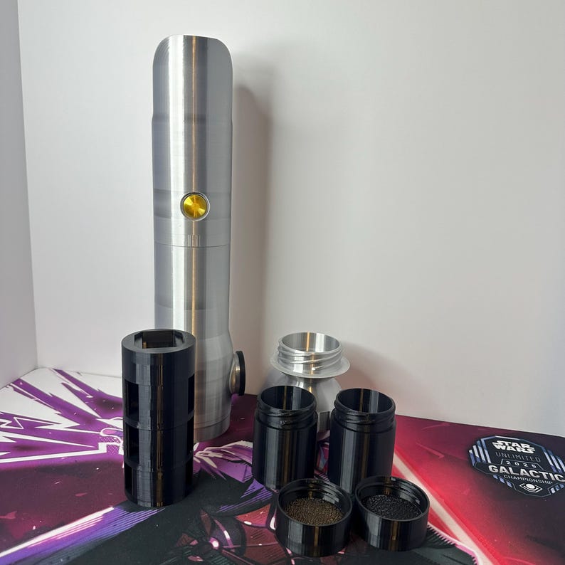 K&ouml;nnte beinhalten: Ein zylindrischer Griff aus silbernem Metall mit einem goldenen runden Knopf, begleitet von mehreren schwarzen Kunststoffteilen, darunter eine hohle H&uuml;lse, zwei kleine Beh&auml;lter, zwei passende Deckel und ein silbernes Metallverbindungsst&uuml;ck.