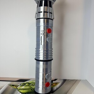 K&ouml;nnte beinhalten: Ein silbernes Lichtschwert-Requisit mit detailliertem Design, das einen zylindrischen K&ouml;rper mit roten und grauen Kn&ouml;pfen aufweist. Der obere Teil hat ein geripptes Design. Das Requisit ist etwa 28 cm hoch.