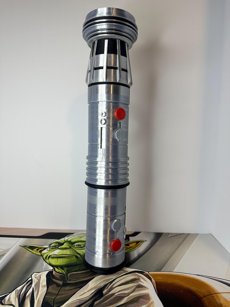 K&ouml;nnte beinhalten: Ein silbernes Lichtschwert-Requisit mit detailliertem Design, das einen zylindrischen K&ouml;rper mit roten und grauen Kn&ouml;pfen aufweist. Der obere Teil hat ein geripptes Design. Das Requisit ist etwa 28 cm hoch.