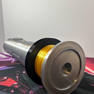 May include: A cylindrical prop with a metallic silver finish, featuring a prominent black circular guard and a bright yellow ribbed section. The front end is capped with a flat, textured silver disc containing a central circular opening.