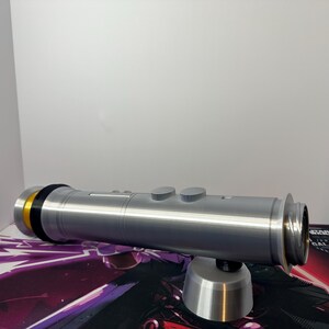 May include: A silver-colored cylindrical lightsaber hilt featuring a gold-toned accent ring at the base, a black spacer, and two circular buttons on the top surface. The hilt is supported by a small, matching silver-colored conical stand.