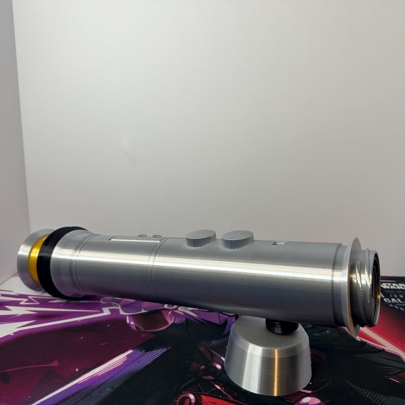 May include: A silver-colored cylindrical lightsaber hilt featuring a gold-toned accent ring at the base, a black spacer, and two circular buttons on the top surface. The hilt is supported by a small, matching silver-colored conical stand.