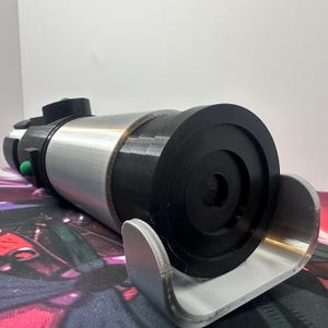 K&ouml;nnte beinhalten: Ein zylindrisches Requisit mit silberner Metallic-Oberfl&auml;che und schwarzen Kunststoffkomponenten. Das Objekt verf&uuml;gt &uuml;ber eine runde schwarze Endkappe, einen silbernen Mittelk&ouml;rper und einen schwarzen Griffbereich mit kleinen gr&uuml;nen Kn&ouml;pfen.