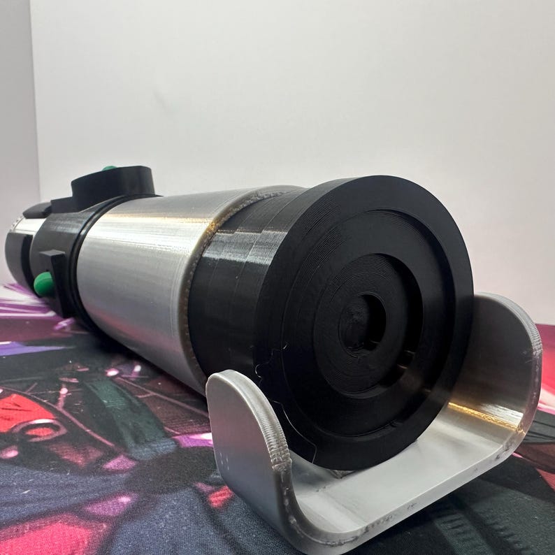 K&ouml;nnte beinhalten: Ein zylindrisches Requisit mit silberner Metallic-Oberfl&auml;che und schwarzen Kunststoffkomponenten. Das Objekt verf&uuml;gt &uuml;ber eine runde schwarze Endkappe, einen silbernen Mittelk&ouml;rper und einen schwarzen Griffbereich mit kleinen gr&uuml;nen Kn&ouml;pfen.