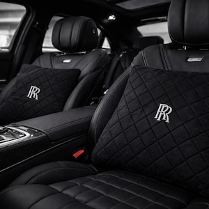 Könnte beinhalten: Schwarze, gesteppte Autositzkissen mit einem weiß gestickten "R"-Logo. Die Kissen befinden sich auf schwarzen Ledersitzen in einem Luxusfahrzeug. Der Innenraum ist dunkel und elegant, mit Fokus auf Komfort und Stil.