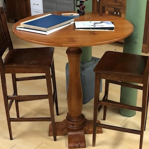 May include: A round, wooden table with a pedestal base, accompanied by two matching wooden chairs. The table has a dark finish and has books on top. The chairs have a ladder-back design. The table is approximately 30 inches in diameter.