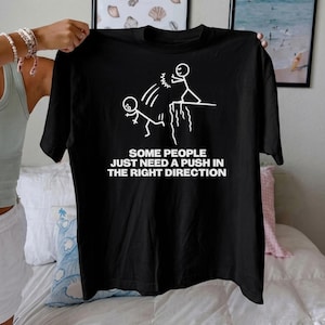 Puede incluir: Camiseta negra con un gráfico blanco de figuras de palo, una empujando a otra por un acantilado. El texto "SOME PEOPLE JUST NEED A PUSH IN THE RIGHT DIRECTION" está debajo. La camiseta está sujeta por una persona.