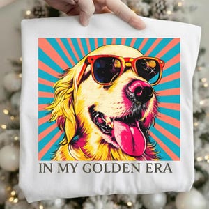 Könnte beinhalten: Ein weißes T-Shirt mit einer Grafik eines Golden Retrievers, der eine rote Sonnenbrille trägt. Der Hund ist vor einem Hintergrund aus blaugrünen und korallenfarbenen Sonnenstrahlen abgebildet. Darunter steht der Text IN MY GOLDEN ERA.