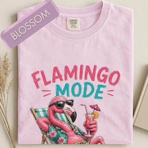 Puede incluir: Una camiseta de manga corta de color rosa claro con el texto "FLAMINGO MODE" en rosa y turquesa. Debajo del texto hay un gráfico de un flamenco rosa con gafas de sol negras, descansando en una silla de playa a rayas con un cóctel en la mano.