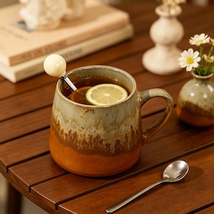 May include: A ceramic mug with a two-tone glaze featuring a light gray top and a warm orange-brown base. The mug contains a dark liquid with a floating lime slice and a metal stirring spoon with a round white handle. A separate silver spoon lies nearby.