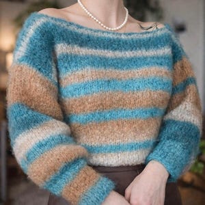 Könnte beinhalten: Ein blau, beige und weiß gestreifter Pullover mit einem Off-Shoulder-Ausschnitt. Der langärmlige Pullover ist aus weichem, strukturiertem Garn gefertigt. Die Farbpalette umfasst Blau-, Beige- und Weißtöne und sorgt für einen gemütlichen und stilvollen Look.