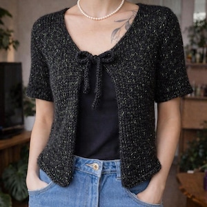 Könnte beinhalten: Dunkelgraue Strickjacke mit kurzen Ärmeln und V-Ausschnitt. Die Strickjacke hat einen Bindverschluss an der Brust und ein dezentes gesprenkeltes Muster. Das Model trägt eine blaue Jeans und ein schwarzes Top.