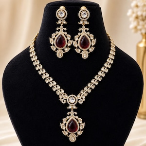 Puede incluir: Un conjunto de collar y pendientes dorados sobre un maniquí negro. El collar y los pendientes presentan piedras preciosas claras, redondas y en forma de lágrima, con una gran piedra roja oscura en forma de lágrima en el centro.