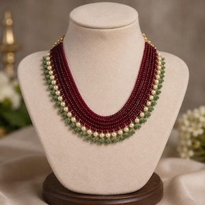 May include: A multi-strand necklace featuring deep red beads arranged in several layers. The necklace is accented with a row of white pearls and small green beads along the bottom edge, creating a layered design with a structured, curved shape.