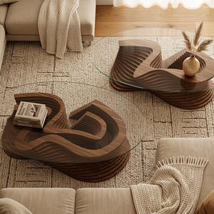 May include: Two modern coffee tables with a unique, layered wood design and clear glass tops. One table has a stack of books on it, and the other has a small vase with decorative grasses. The tables sit on a neutral-toned rug.