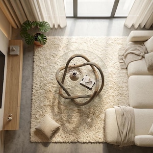 Op de afbeelding: Een bovenaanzicht van een moderne woonkamer met een crèmekleurige hoekbank, een shaggy vloerkleed en een unieke glazen salontafel met een geweven basis. Een plant in pot en sierkussens geven de ruimte warmte.