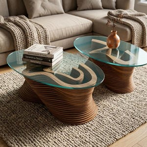 May include: Two oval coffee tables with clear glass tops and wooden bases. The bases have a unique, layered design. One table has a stack of books on top. The tables sit on a woven rug in a living room setting.