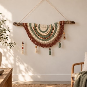 Peut inclure: Une décoration murale en macramé composée de cordes couleur crème, vert sauge et terre cuite, suspendue à une branche de bois naturel. L'objet présente des couches semi-circulaires concentriques, des tresses, des perles et des pompons.