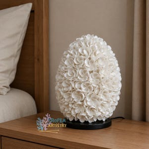 May include: A white, egg-shaped lamp covered in small, shell-like elements. The lamp sits on a black base and is plugged into an outlet. The lamp is on a wooden nightstand next to a bed.