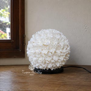 Könnte beinhalten: Eine kugelförmige weiße Lampe aus kleinen, muschelartigen Elementen, die auf einem schwarzen Sockel steht. Die Lampe hat ein Netzkabel und steht auf einer Holzoberfläche. Der Text "Tropea Artistry BALI" ist sichtbar.