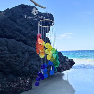 May include: A circular wind chime featuring numerous translucent, multicoloured discs in a spectrum of red, orange, yellow, green, and blue. The discs are suspended by thin strings from a circular wooden frame. The text Tropea Artistry BALI is visible.