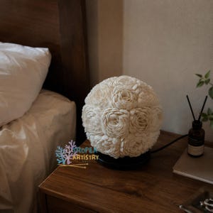 May include: A cream-colored, spherical lamp with a floral design, resting on a black base. The lamp is on a wooden nightstand next to a bed. The lamp's design features multiple flower shapes. The text "ROPEA ARTISTRY" is visible on the nightstand.