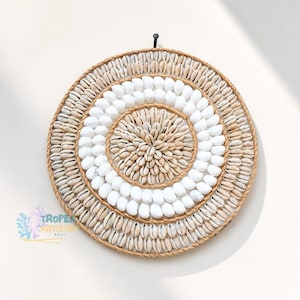 Könnte beinhalten: Runde Wanddekoration mit einem Spiralmuster aus Muscheln und weißen Steinen. Der äußere Rand ist mit einem Naturseil eingefasst. Das Kunstwerk ist an einer weißen Wand befestigt und schafft eine Küstenatmosphäre.