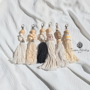 Puede incluir: Seis llaveros decorativos con cierres de mosquetón plateados, cuentas de madera natural y varias conchas, incluyendo cauríes y conos. Las borlas son de cordón de algodón en crema, beige y negro. El texto dice Tropea Artistry BALI.