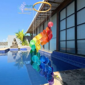 May include: A colorful wind chime with a rainbow gradient of circular, iridescent shells. The shells hang from a gold-colored ring, creating a decorative and eye-catching outdoor accent. The wind chime is suspended near a pool.