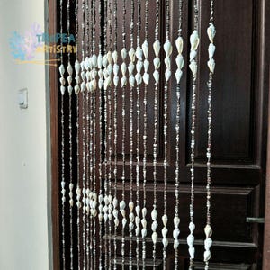 Pode incluir: Uma cortina de contas com múltiplos fios verticais de corda castanho-escura adornados com pequenas conchas brancas em forma de cone. Os fios pendem em frente a uma porta de madeira castanho-escura. O texto TROPEA ARTISTRY é visível no canto superior esquerdo.