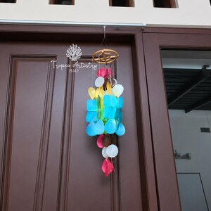 May include: A hanging wind chime featuring a circular wooden frame with multiple strands of translucent, colourful shell-like discs in shades of blue, yellow, white, brown, and pink. The text Tropea Artistry BALI is visible in white above the chime.