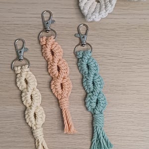 May include: Three macrame keychains featuring a knotted cord design in cream, peach, and sage green. Each keychain is attached to a silver-toned metal lobster clasp. The cords are woven into a series of open loops, ending in a short, frayed tassel.