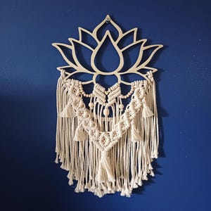 Peut inclure: Une décoration murale composée d'une découpe de fleur de lotus en bois clair sur le dessus, avec une corde en macramé blanc cassé nouée en motif géométrique. L'ensemble comprend des perles en bois et plusieurs pompons suspendus.