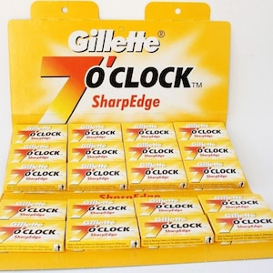 May include: A yellow retail display card holding multiple small rectangular boxes of Gillette 7 O'CLOCK SharpEdge razor blades. Each box features the brand name and product text in black and red against a bright yellow background.