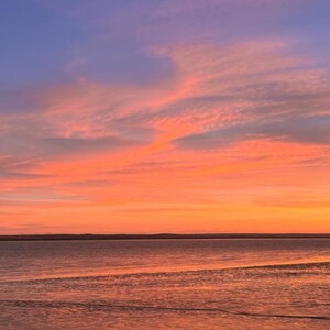 Könnte beinhalten: Ein lebhafter Sonnenuntergang über einem ruhigen Gewässer mit einem Farbverlauf am Himmel, der von tiefem Blau oben zu leuchtenden Orange- und Rosatönen am Horizont übergeht, wobei sich die Farben sanft auf der Wasseroberfläche spiegeln.