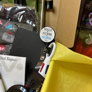 Peut inclure: Un enrouleur de badge rond portant le texte MALE + NURSE = MURSE en lettres noires et bleues avec une ic&ocirc;ne de st&eacute;thoscope. L'enrouleur est fix&eacute; &agrave; un stylo r&eacute;tractable blanc avec des boutons d'encre rouge et bleue, pos&eacute; dans une bo&icirc;te en papier de soie jaune.