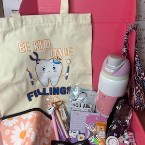 Peut inclure: Une bo&icirc;te cadeau rose contenant un sac fourre-tout cr&egrave;me avec un graphique de dent et le texte BE KIND I HAVE FILLINGS TOO, une gourde rose et blanche, une pochette &agrave; motifs marguerites, des stylos, un bloc-notes, de la cr&egrave;me pour les mains.