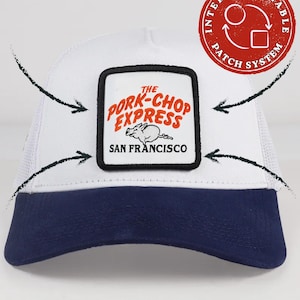 Op de afbeelding: Een witte truckerpet met een donkerblauwe klep en een vierkante patch aan de voorzijde. De patch bevat de tekst THE PORK-CHOP EXPRESS SAN FRANCISCO en een illustratie van een varken. Een rode cirkelvormige afbeelding duidt op een verwisselbaar patchsysteem.