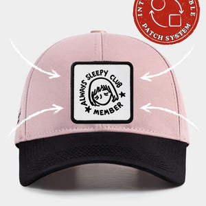 May include: A light pink baseball cap with a black brim and a square white patch on the front. The patch features a black line drawing of a face with closed eyes and the text Always Sleepy Club Member. A red circular logo indicates an interchangeable patch system.