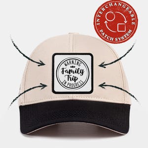May include: A beige baseball cap with a black brim and a square patch on the front. The patch features a circular design with the text "WARNING Family Trip IN PROGRESS" and three small stars. A red circular icon indicates an interchangeable patch system.