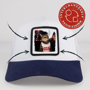 Op de afbeelding: Een witte en marineblauwe truckerpet met een vierkante patch met een afbeelding van een kat in een wit hemdje met de tekst USA in het rood gedrukt. De kat staat afgebeeld tegen een achtergrond van vuurwerk en een Amerikaanse vlag. De patch is afneembaar.