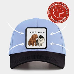 May include: A pale blue baseball cap with a black peak. The cap has a square patch with the text "MISO SUSHI" and illustrations of a dog and a cat. A red circular patch with the text "INTERCHANGEABLE PATCH SYSTEM" is also visible.