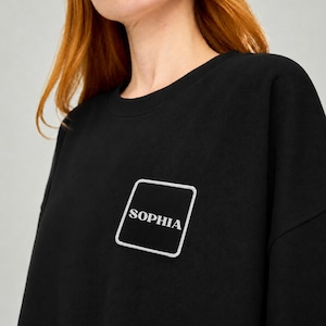 May include: A black long-sleeve sweatshirt featuring a square patch on the chest. The patch is black with a white border and contains the white text SOPHIA in a serif font. The garment has a crew neckline and a simple, clean design throughout.
