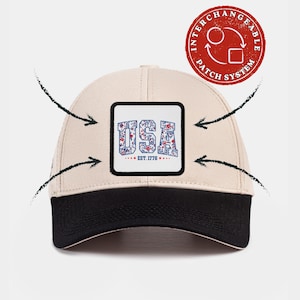 May include: A beige baseball cap with a black brim and a square patch on the front. The patch features the text USA in a blue and red floral pattern, with the text EST. 1776 below it. A red circular logo indicates an interchangeable patch system.