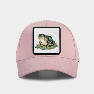 May include: A pale pink baseball cap with a square patch depicting a detailed illustration of a green and brown frog. The frog is on a patch of green grass and rocks. The cap has a curved peak and a classic six-panel design.