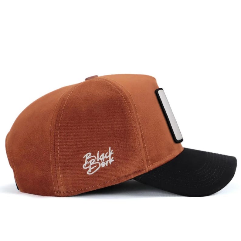 May include: A rust-colored baseball cap with a black brim and a rectangular patch on the side. The side panel features white embroidered text that reads Black B&oslash;rk. The cap has a structured crown and a curved visor, showcasing a two-tone design.